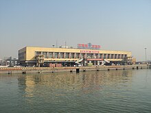 a long ochre low-rise building with broad windows. A pier extend parallel to the building