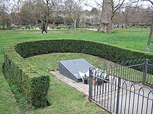 U-shaped hedge enclosure surrounding an oblong slate plaque on a wedge-shaped plinth, with flowers laid at the bottom