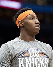 Profile photo of a man wearing a grey “Los Knicks” t-shirt and orange headband looking up to right