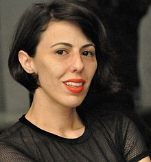 Ortal Ben Dayan, 3/4 angle, seated, wearing a black short-sleeved outfit. Dark hair in a bob hairstyle, and wearing orange-red lipstick.