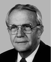 A man with gray hair and glasses wearing a black jacket and tie and white shirt