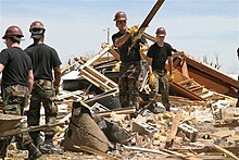 Several uniformed men are tossing debris from a pile of rubble that was left of the Greensburg High School. It is daytime, and the men seem to be having a conversation with each other.