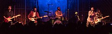 Four men performing on stage. From right to left: man in a dark shirt with an electric guitar, man in a red flannel with an electric bass guitar, man in a tan shirt behind a drum kit, man in a pink shirt with a microphone in his hand, and a man black and white shirt with an electric guitar.