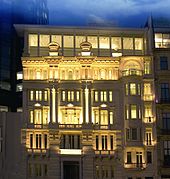 A six-story white building with light emanating from its windows under cloudy evening sky