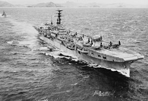 A small aircraft carrier travelling at speed through choppy water. Several propeller aircraft with folded wings are sitting on the flight deck. A small warship is following in the carrier's wake, and a rocky, hilly shoreline can be seen across the horizon