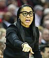 A woman in black glasses and a black shirt stands on the basketball court, coaching her team