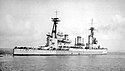 Right rear oblique shot of a large gray warship with two masts, three funnels, and two gun turrets visible. A number of people are visible on her decks.