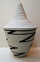 Photograph depicting a bowl shaped off-white woven basket with tall conical lid and black zig-zag pattern