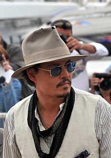 A moustached man wearing a hat, sunglasses, a striped shirt, a white jacket and a scarf. Three photographers and two boats can be seen behind him.