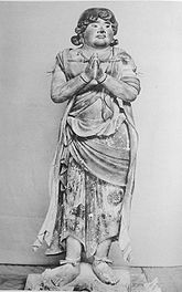 Front view of a standing statue with plump face. His hands are joined in front of his breast with palms facing each other as if praying. He is barefoot and some painted patterns are visible on his cloths. Black and white photograph.