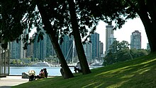 A green grassy hill dotted with trees slopes down to a paved area with benches. Beyond lies water, docks, and a yacht, and skyscrapers.