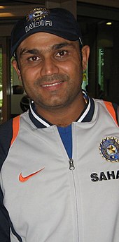 Virender Sehwag wearing a grey jacket and a cap, smiling.