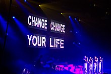 A large stage screen picturing the title 'Change Your Life' in capital font. Below the screen are four women standing on a stage as blue and pink stage lighting shines upon them.