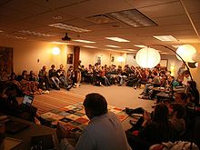 People seated on a circle of chairs in a large room with hanging lights from the ceiling and a table visible in the foreground