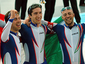 Head and torso photo of three smiling men, wearing glasses on their heads and blue and white jumpsuits with Italia written on the chest. The man in the middle is holding a big Italian flag behind him, while the other two are embracing him.