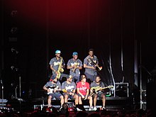 A group of seven men sit and stand on a stage against a dark backdrop.