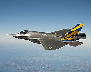 Gray fighter aircraft soars in a clear blue sky with sea coast below.