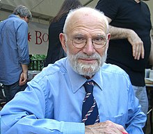 A grey-haired Oliver Sacks with glasses, a beard and a blue shirt with three people in the background