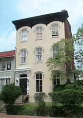 A yellow three story town house with steps in front and a small lawn with shrubs and trees.