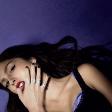 Olivia Rodrigo in a black dress biting her thumb on a dark purple floor