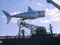 A large model shark is hoisted by a crane as two men watch it.