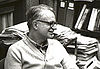 man wearing a sweater and glasses in a clutttered office against a background of papers and bookshelves