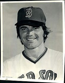 A black-and-white photo of a man's head and chest. He is wearing a baseball cap with the letter "B" and a white baseball jersey that says "RED SOX", partially obscured.