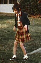 A slightly grainy color picture of Genie, facing slightly right of the camera, walking by herself outside in the Children's Hospital recreation yard. She is wearing a plaid-patterned dress and thin sweater and looks extremely pale, emaciated, and expressionless. Her limbs are exposed and look extremely thin. Both of her knees are very bent, and her arms are bent forward with both hands hanging down as she holds them out in front of her.