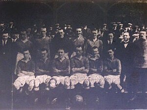 Six footballers are sitting on a bench, five more are standing up behind them. A number of men in flat caps are standing in the background.