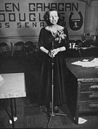 A well-dressed, tired looking woman stands before a microphone, part of a campaign banner "Helen Gahagan Douglas" is visible in the background.